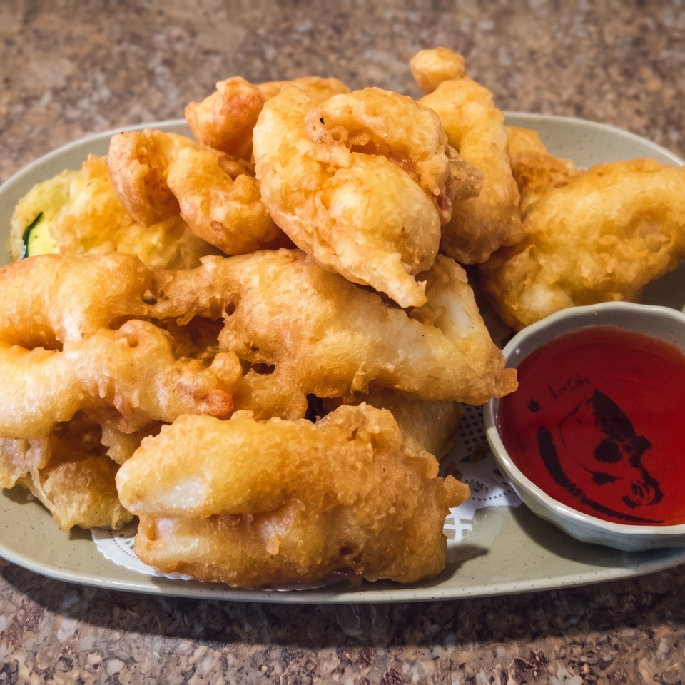 Plate of golden battered fried seafood served with a small bowl of red dipping sauce on a stone countertop.